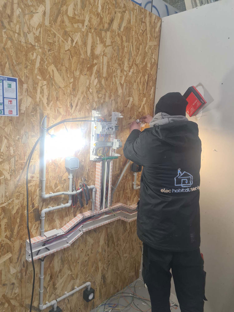 Technicien Elec Habitat Service installant un coffret de disjoncteurs neuf sur une cloison en cours de chantier